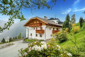 A white building with wooden balconies and a stone foundation, surrounded by greenery and mountains.