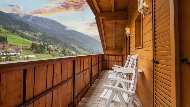 A wooden balcony with white plastic chairs and a table, overlooking a mountainous landscape.