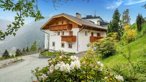 A white building with wooden balconies and a stone foundation, surrounded by greenery and mountains.