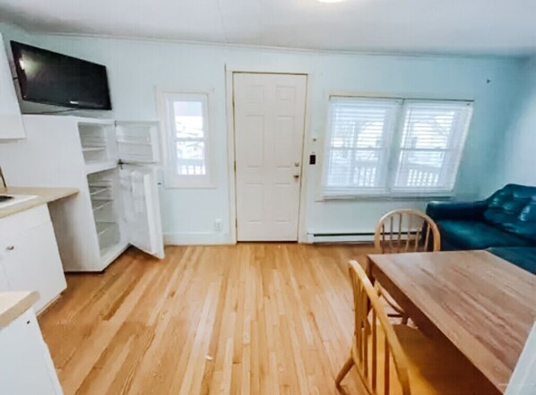 A room with a wooden floor, a dining table with chairs, a television mounted on the wall, and a window with blinds.