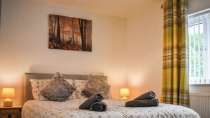 Ein Schlafzimmer mit einem großen Bett, zwei Nachttischen, einem Waldgemälde und einem Fenster mit Vorhängen.