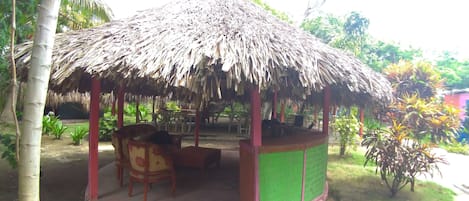 A thatched-roof gazebo with a table and chairs, surrounded by greenery.