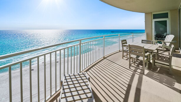 A balcony with a view of the ocean and a pier. There are chairs and a table on the balcony.