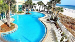 A swimming pool with a curved design, surrounded by palm trees and lounge chairs, with a beach and ocean view.
