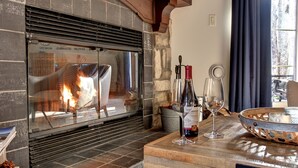 Una chimenea de piedra con puerta de vidrio, una mesa de madera con una copa y una botella de vino, y una ventana con cortinas.