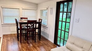 A dining area with a wooden table and chairs, a beige sofa, and large windows with blinds.