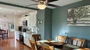 A dining area with a wooden table and chairs, a kitchen in the background, and a ceiling fan.