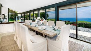 A modern dining area with a large table, white chairs, and a view of the ocean.