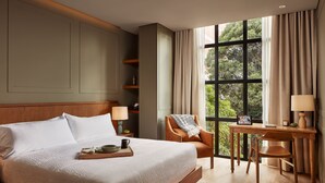 A hotel room with a large bed, a desk with a chair, a window with curtains, and a view of greenery outside. - Hotel Coco (Bogotá)