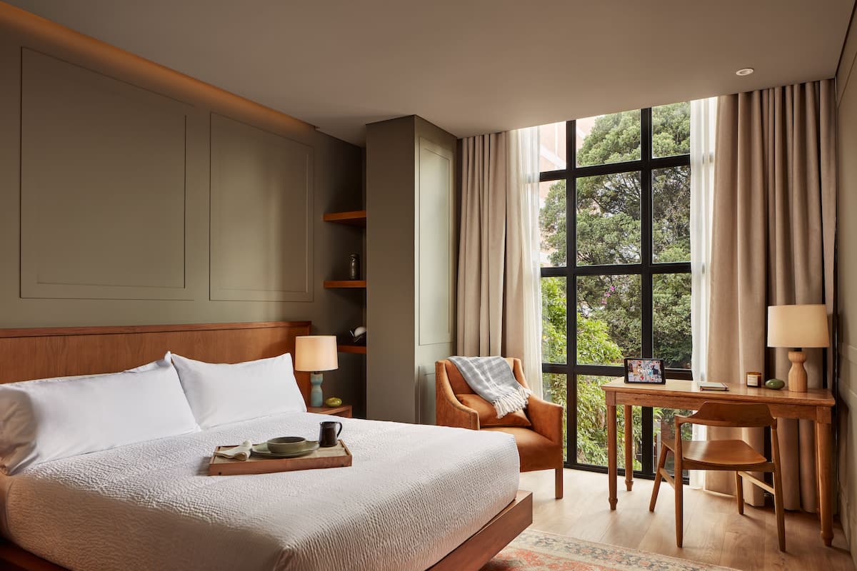 Quarto de hotel com uma cama grande, uma mesa com cadeira, janela com cortinas e vista para o verde lá fora.