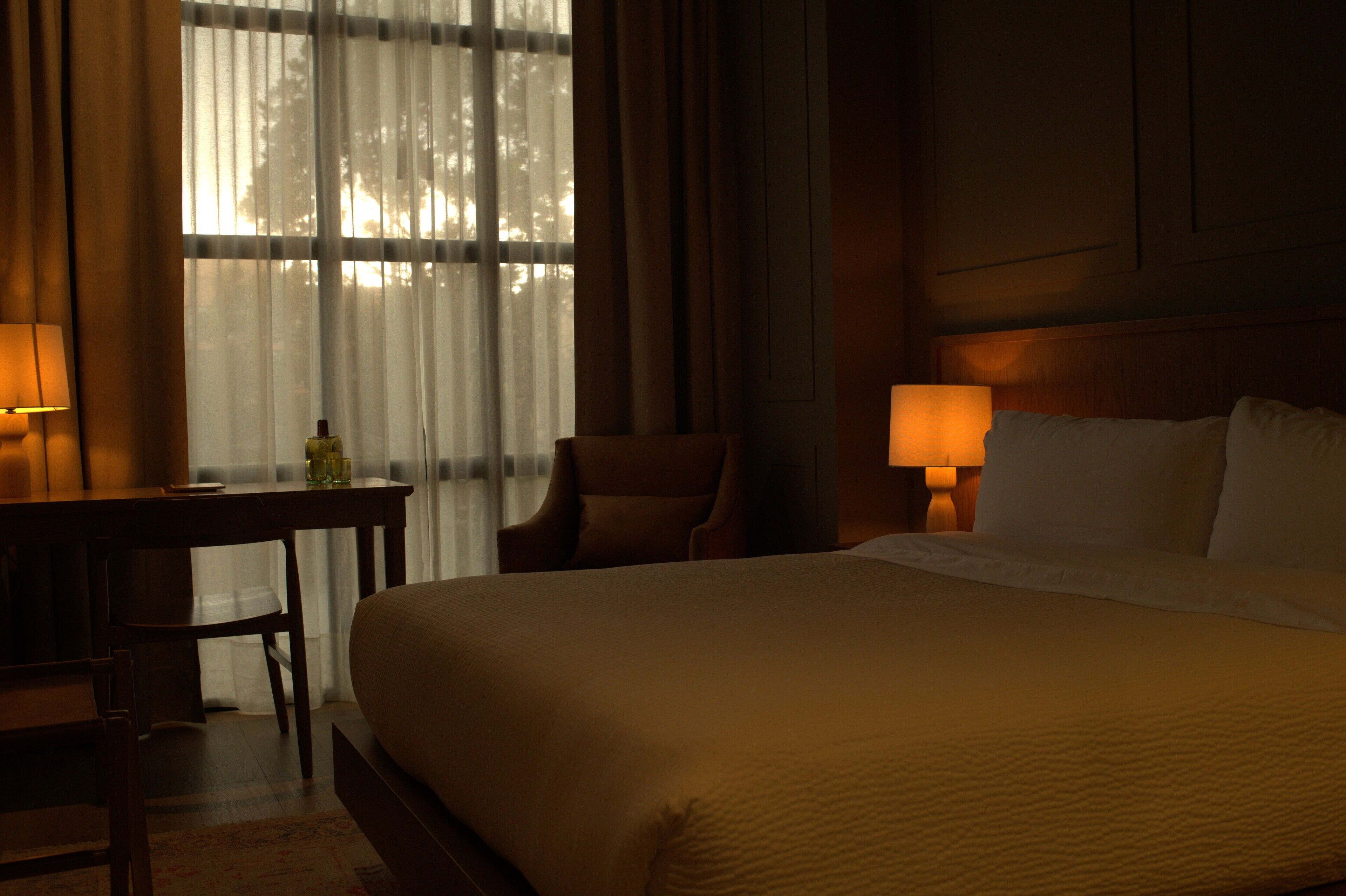 Una habitación de hotel con una cama grande, dos lámparas de noche, un escritorio con una silla y una ventana con cortinas ligeras.