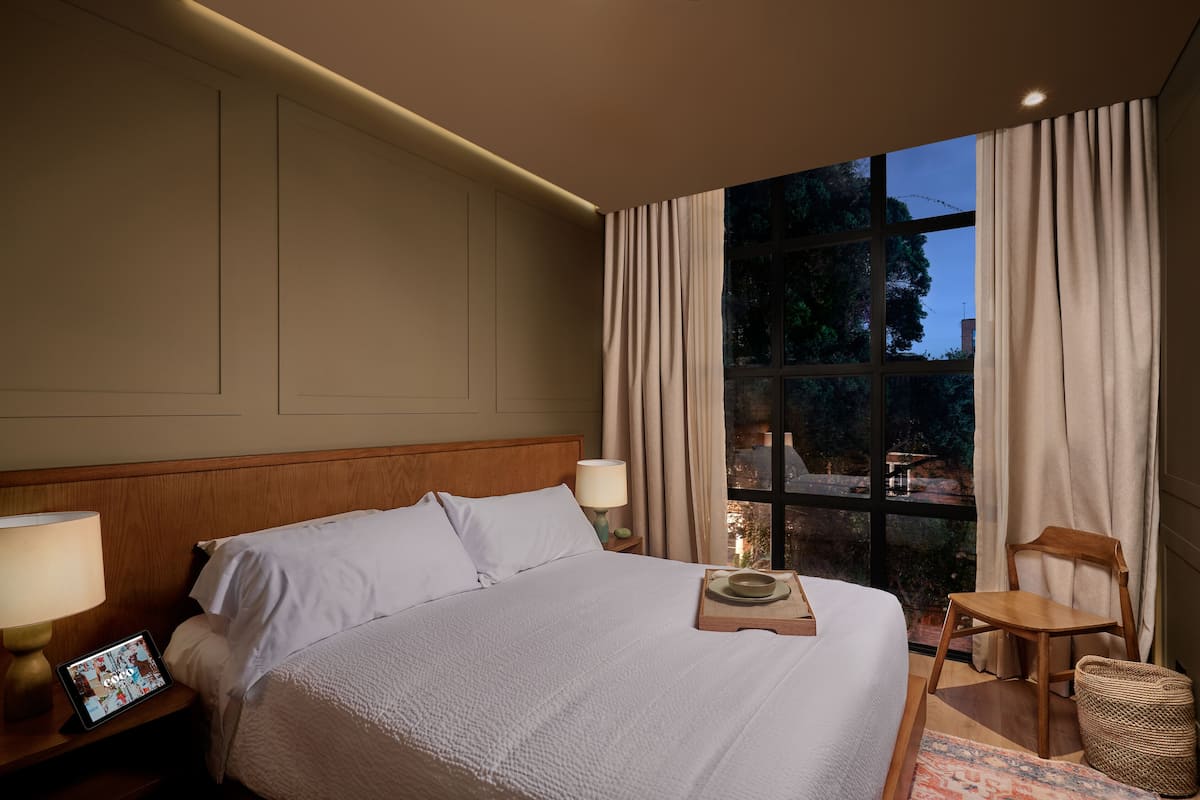 Quarto de hotel com uma cama grande, mesa de cabeceira com abajur, uma cadeira e vista para o exterior pela janela.