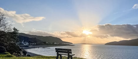 A bench by a lake with a sunset view, grassy area, and distant hills.