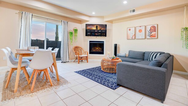 A modern living room with a grey sofa, a dining table with chairs, a fireplace, and a TV mounted on the wall.