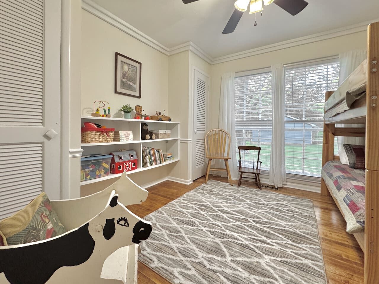 A cozy room with a wooden floor, a patterned rug, a bookshelf, a chair, and a bunk bed.