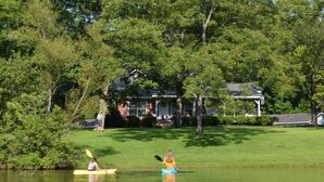 A calm lake with kayaks, surrounded by lush greenery and a house in the background. - Pond House on the Plains, Auburn, Firetruck rides (Opelika)