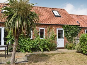 A brick house with a red-tiled roof, white doors, and windows, surrounded by greenery and a palm tree.