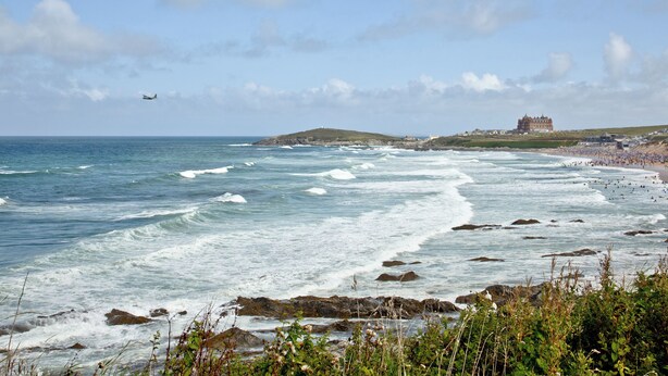 A coastal landscape with waves, a beach, and a large building in the distance. - Apartment 1 Waterrs Edge (Newquay)