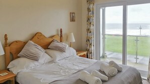 A neatly made bed with pillows and rolled towels, a bedside lamp, and a view of the sea through a large window. - Apartment 1 Waterrs Edge (Newquay)