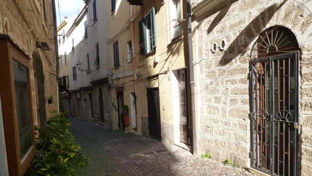 A narrow cobblestone street flanked by historic buildings with arched windows and doors, a stone wall with a metal gate, and potted plants. - Loft in the old Town (Alghero)