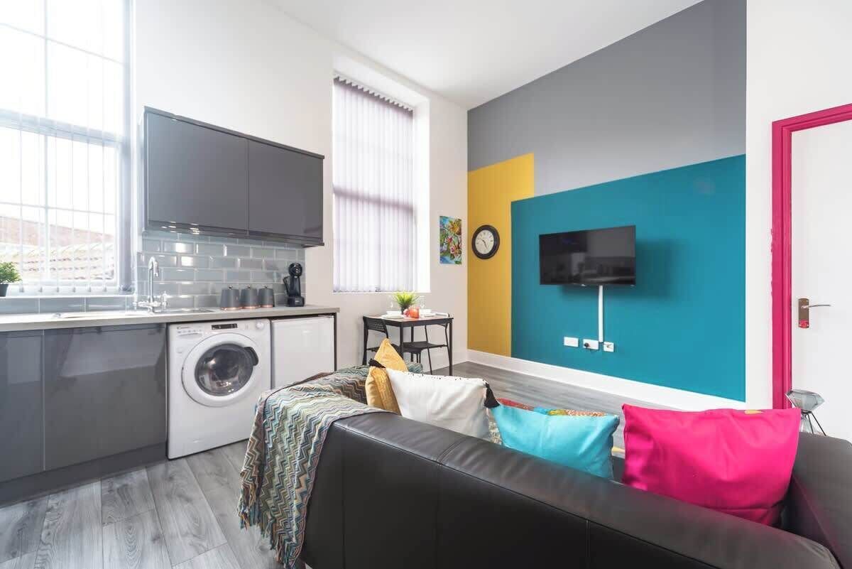 A compact living space with a kitchenette, a washing machine, a flat-screen TV, and a couch with colorful cushions.