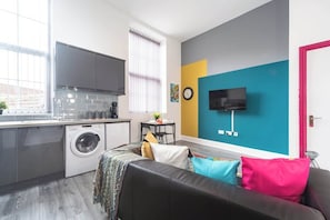 A compact living space with a kitchenette, a washing machine, a flat-screen TV, and a couch with colorful cushions. - Jesouth Exquisite Suite Dazzling Chic Centre (Kingston upon Hull)