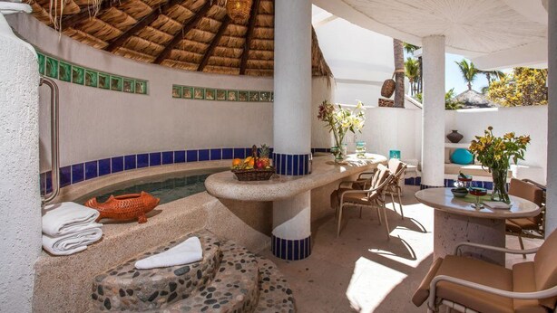A sunlit indoor pool area with a thatched roof, stone seating, and a bar with a fruit bowl. - Beautiful view of the arch, Club Cascadas suite with kitchen. (Cabo San Lucas)