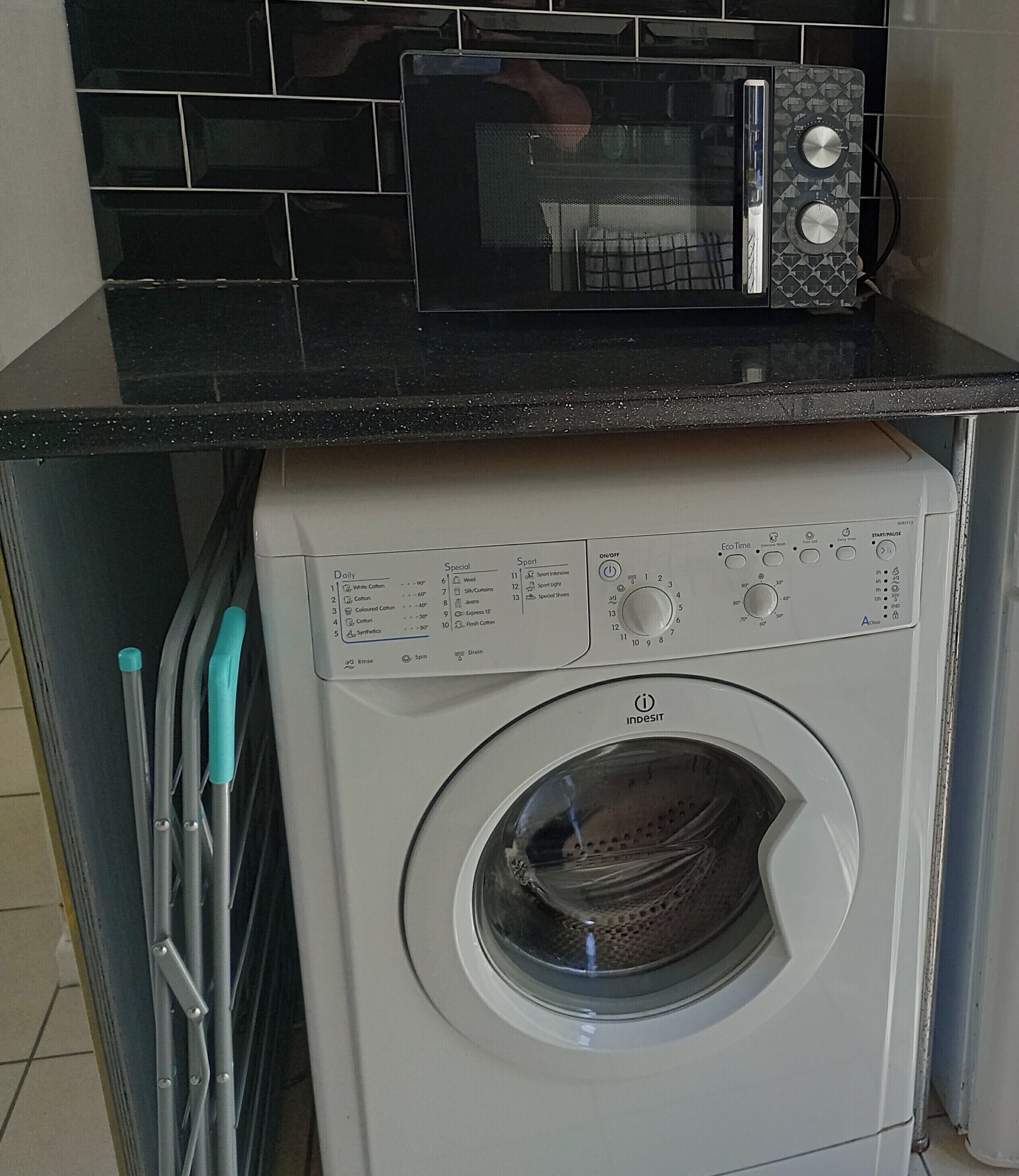 Eine kompakte Waschmaschine und Trockner-Kombination mit Bedienfeld, in einer Küchenecke mit schwarzem Fliesenrückwand und einer Mikrowelle darüber.