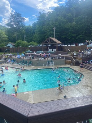 Ein Schwimmbecken mit badenden und entspannten Personen. Sonnenschirme, Liegestühle und ein Holzdeck umgeben das Becken.