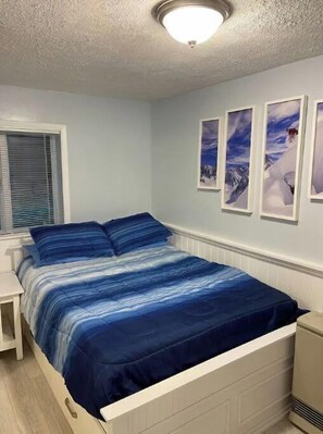 Ein Schlafzimmer mit einem Bett, einem Nachttisch, einem Fenster mit Jalousien und drei gerahmten Bildern an der Wand.