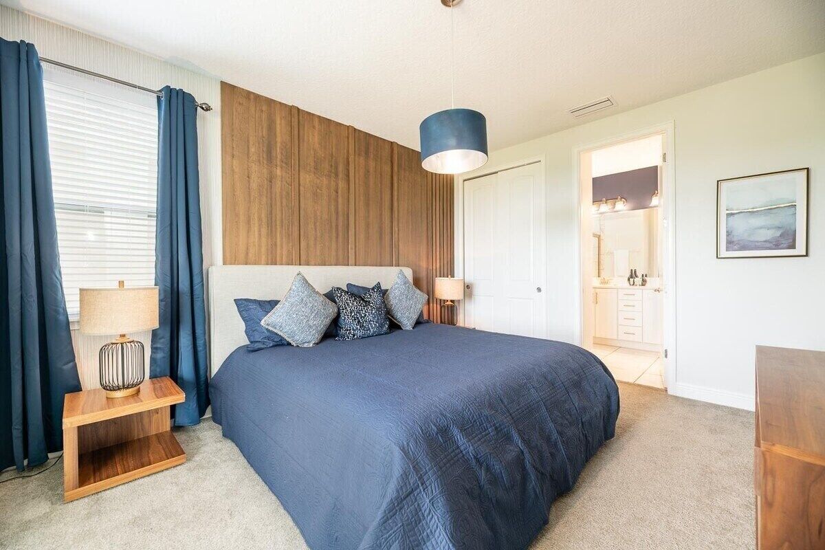 A bedroom with a large bed, a wooden headboard, a bedside table with a lamp, and a view into the bathroom.