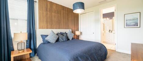 A bedroom with a large bed, a wooden headboard, a bedside table with a lamp, and a view into the bathroom.
