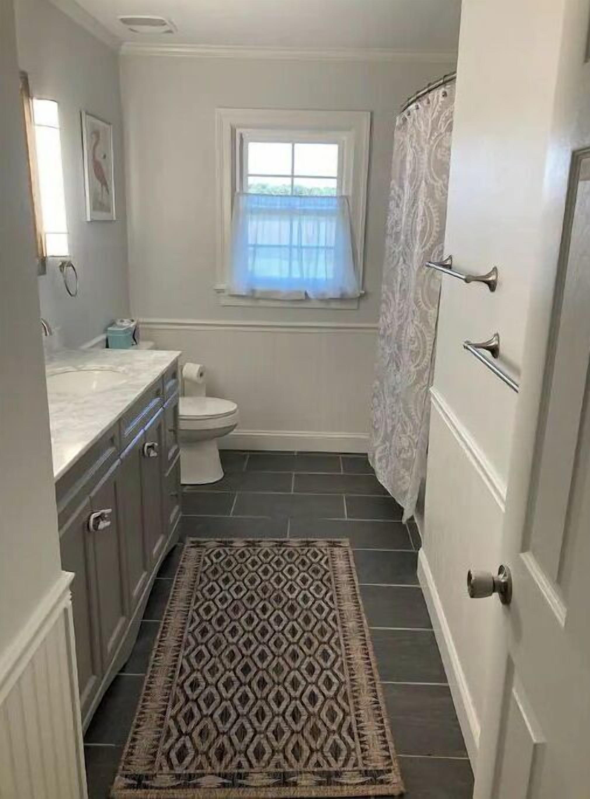 Een kleine badkamer met toilet, een wastafel met handdoekhouder, een gekleurd tapijt en een raam met gordijn.