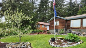 A wooden cabin with a flagpole, surrounded by a well-maintained garden with a lawn, potted plants, and a gravel path.