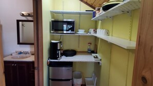 A compact kitchen with a microwave, coffee maker, and a small refrigerator. There are shelves above the counter holding kitchenware.