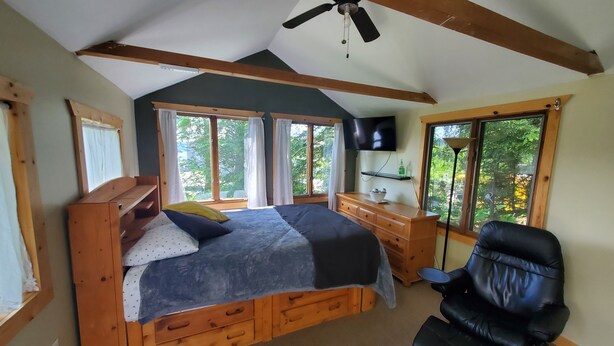A bedroom with a wooden bed, a desk with a chair, a ceiling fan, and large windows offering a view of trees.
