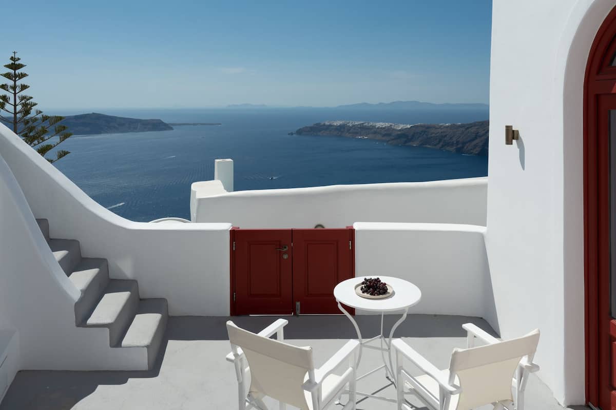 A white balcony with a table and chairs, overlooking a scenic view of the sea and distant islands.