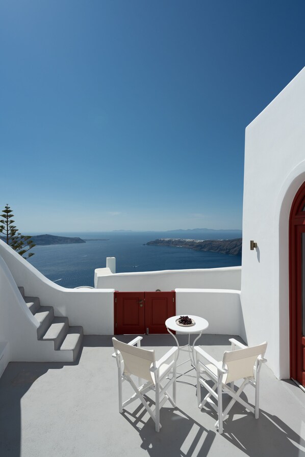 A white balcony with a table and chairs, overlooking a scenic view of the sea and distant islands.