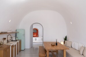 A modern kitchen with a light blue refrigerator, wooden cabinets, and a white countertop. There is a wooden dining table with a bench and a white sofa with cushions. An archway leads to a bathroom with a white sink and an orange accent wall.