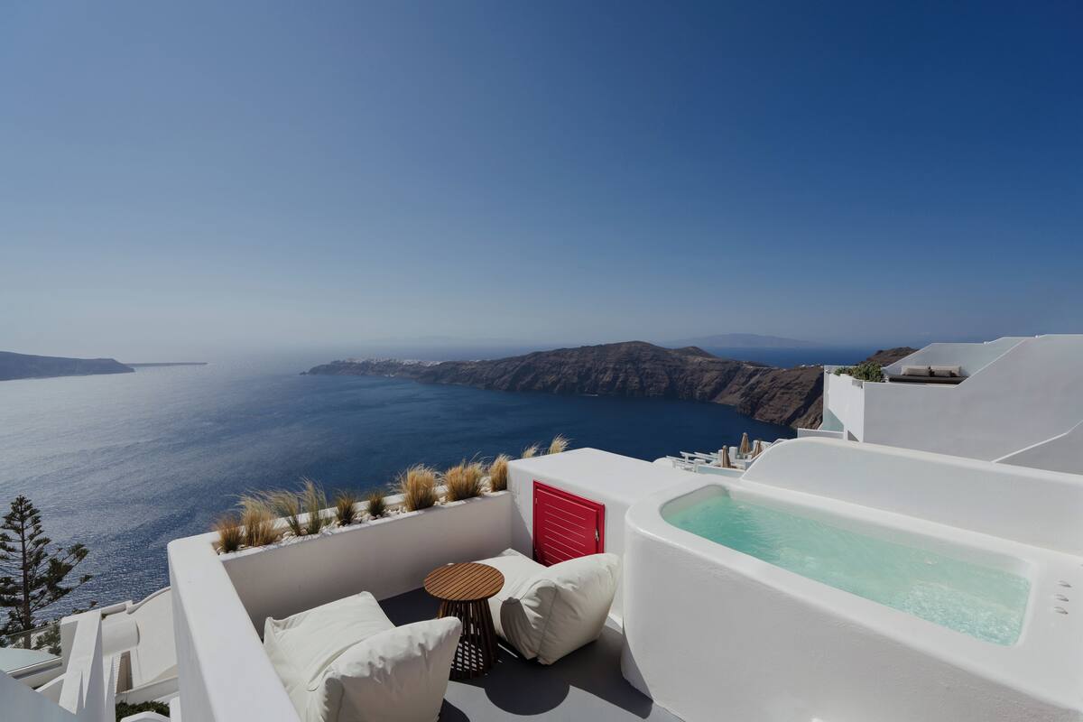 A white infinity pool with a red door, a small round table, and a white chair on a balcony overlooking the sea.