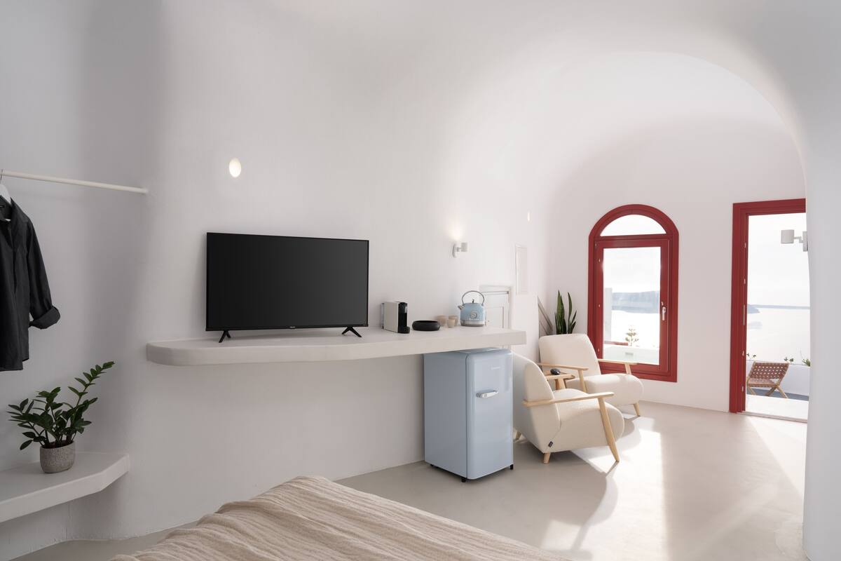 A modern hotel room with a flat-screen TV, a small fridge, a seating area with a chair and a table, and a balcony with a red door.
