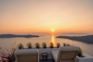 A sunset view from a terrace with two lounge chairs and a small table, overlooking a calm sea and distant landforms.