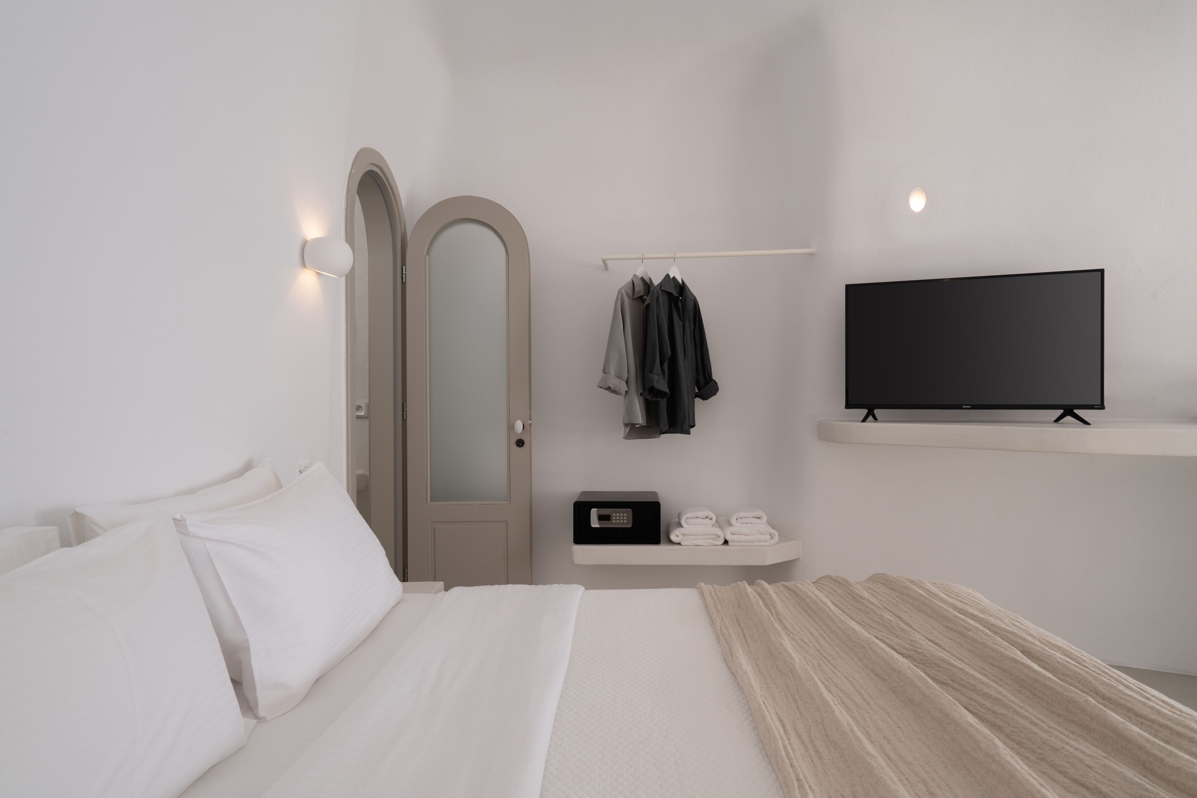 A modern hotel room with a bed, a TV, a wardrobe, and a shelf with towels and a radio.