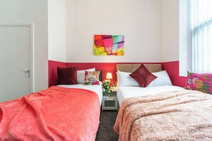 Habitación de hotel con dos camas, un cuadro abstracto colorido, una mesita de noche con lámpara y una ventana con cortinas.