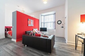 A modern living room with a red accent wall, a black sofa, a flat-screen TV, and a dining area with a table and chairs. - Modern City Centre Apt Near Theatre & Marina (Kingston upon Hull)