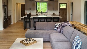 Un ampio soggiorno con un divano modulare grigio, un tavolino in legno e un angolo cottura con pensili in legno scuro e un'ampia finestra.