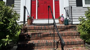 Une maison à deux étages avec une porte d’entrée rouge, des moulures blanches et des volets noirs. Des marches mènent à la porte, et un petit drapeau américain et un pot de fleurs sont placés sur les marches.