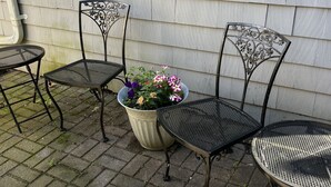 Une terrasse avec deux chaises en métal et une table, un pot de fleurs, et une fenêtre avec des stores.