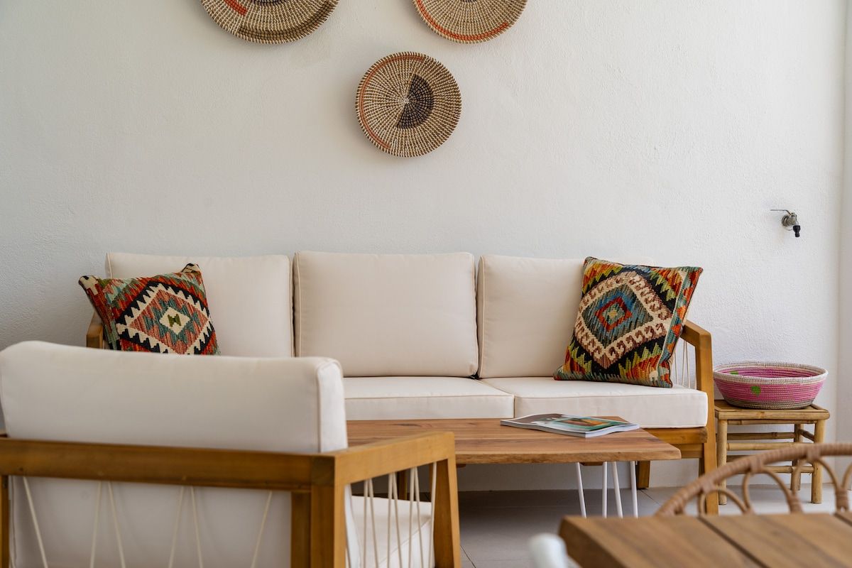 Un salon comprenant un canapé, deux coussins décoratifs à motifs, une table basse en bois et un fauteuil en osier.
