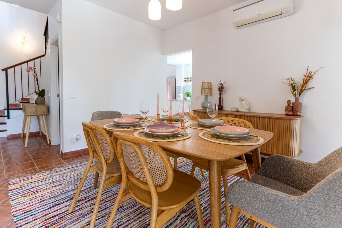 Un coin repas avec une table en bois dressée pour quatre personnes, entourée de chaises en osier, et un tapis rayé aux couleurs vives sur le sol.